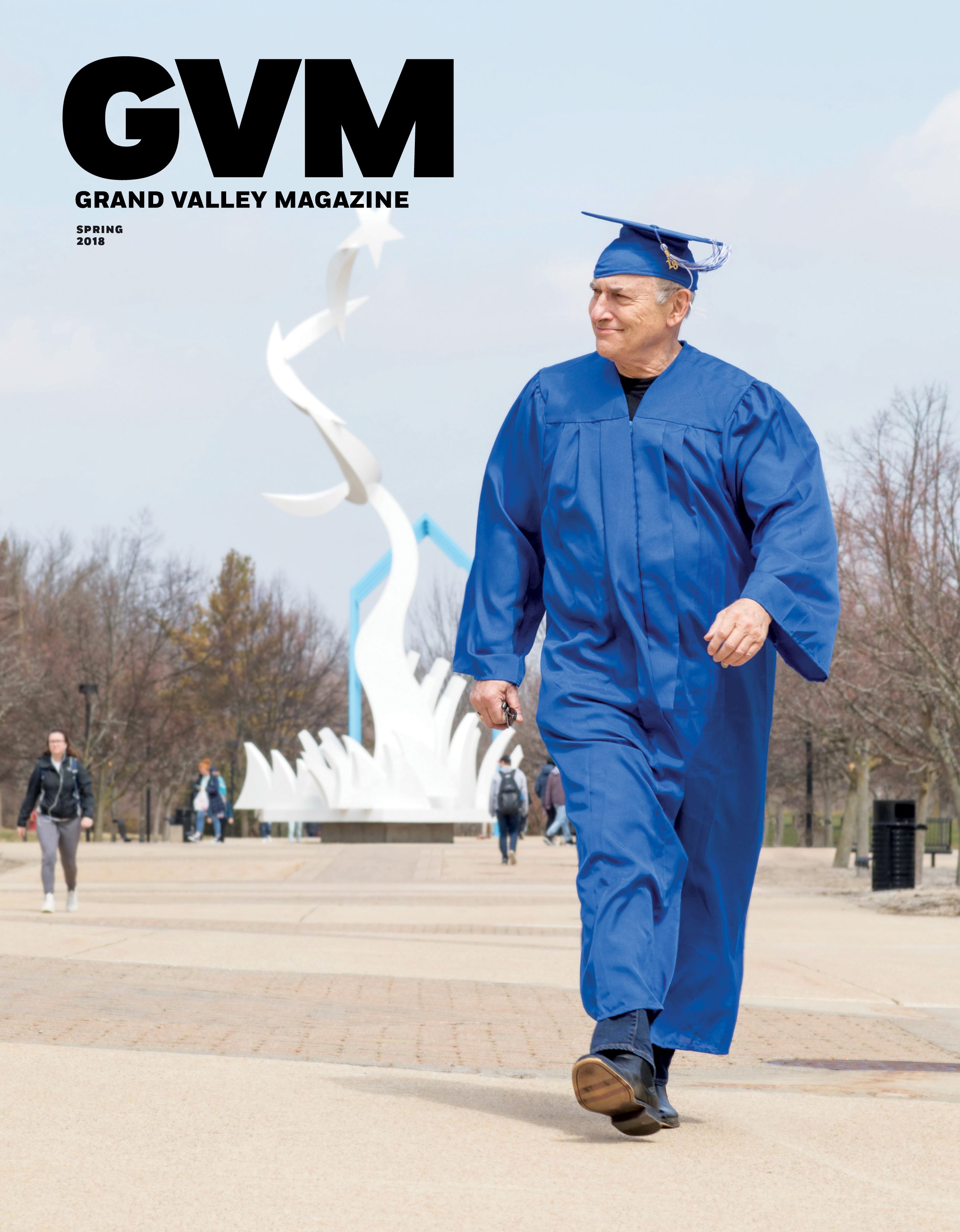an older white gentlemen walking passed the lake hall buildings while wearing a blue cap and gown. the white twisted sculpture is in the background.