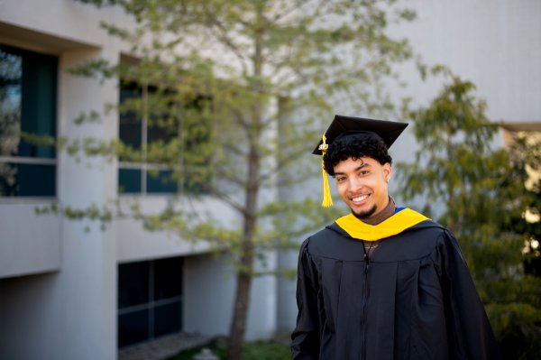 A student wearing a cap and gown with a yellow piece of material draped over the shoulders and a yellow tassel stands in front of a tree and building.