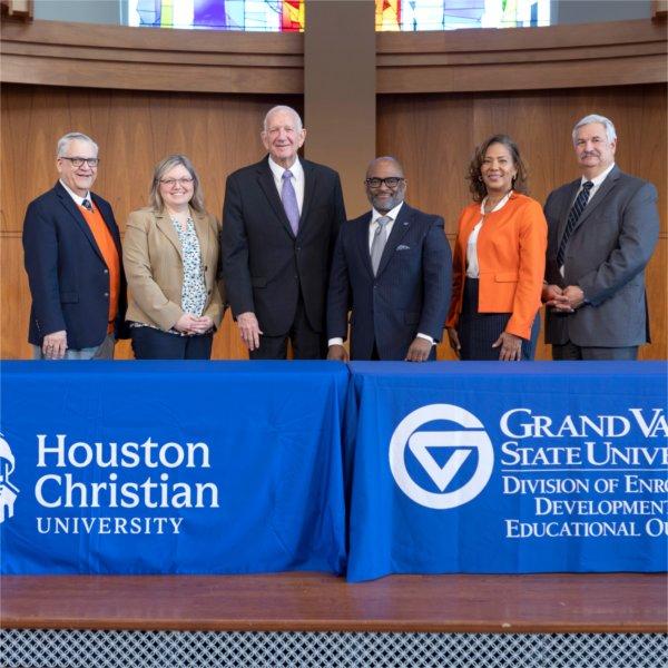 six people standing behind tables with two drapes, one for Houston Christian University, other for GVSU