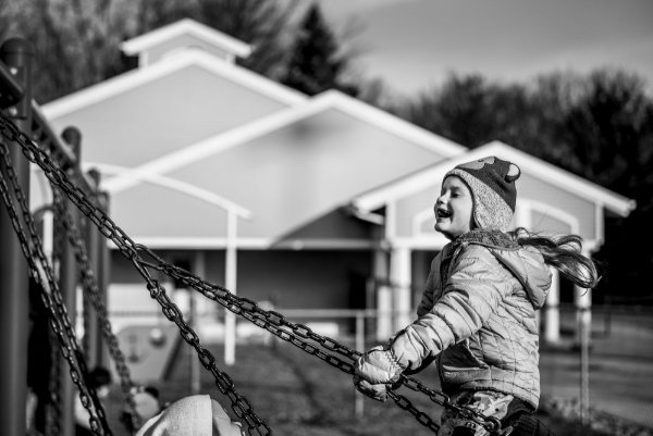 A little girl with a long ponytail and winter hat laughs as she swings on playground equipment outside a preschool.