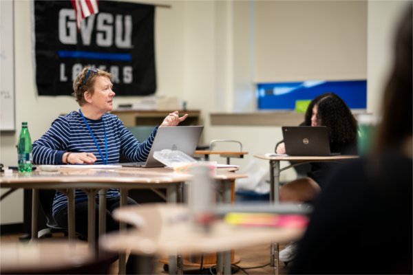 At left, Sarah Cox gestures to a student during a class. A GVSU Lakers flag is tacked to the wall behind them.