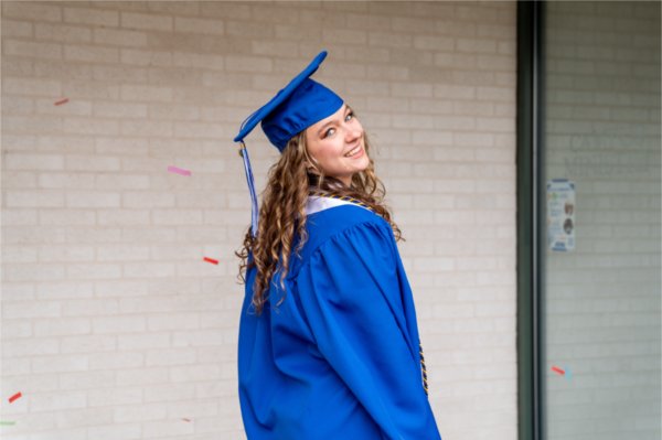 A person wearing a cap and gown smiles while looking over their shoulder. Pieces of red and pink confetti are in the air.