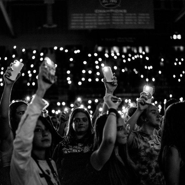 Students hold up their cell phones to give off lights in an arena. Lights can be seen all around.