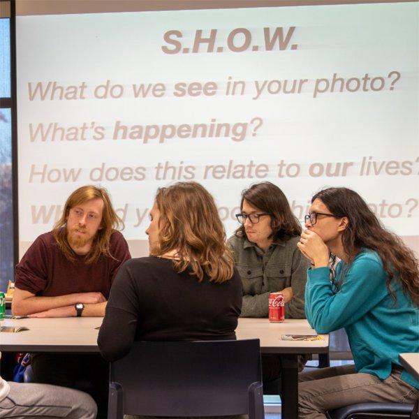 four students at table with projection on screen in background, asking SHOW, what do we see in your photo?