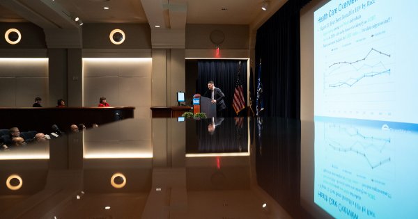 A person wearing a suit stands at a lectern during a talk where they are showing graphs.  