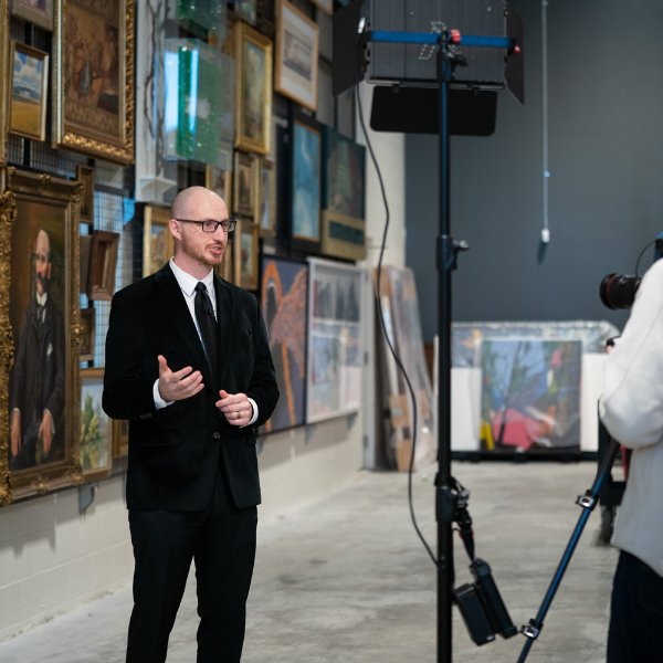 Nathan Kemler is interviewed for a video in a large room with lots of artwork behind him.