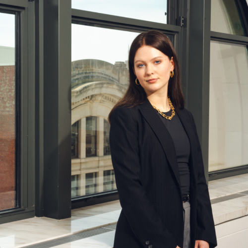 Olivia Nolff standing in black jacket with gold necklace on, against set of windows