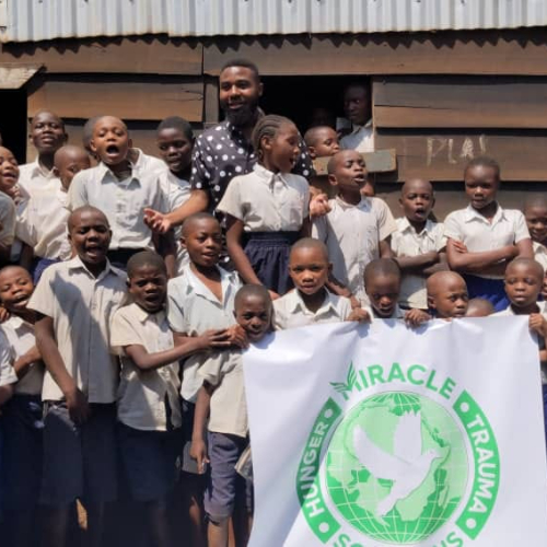 lots of small children in white shirts, Miracle Hunger and Trauma Solutions flag and Kyezie Bwanangela standing in back