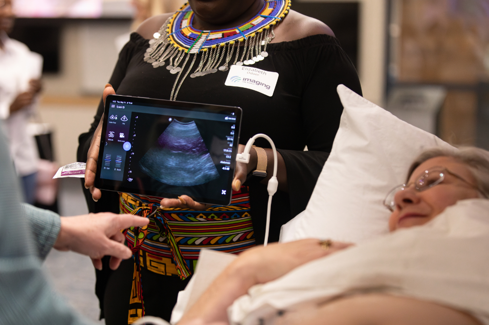 woman portraying patient in bed on right, two others hold an iPad with technology to do an ultrasound on the patient