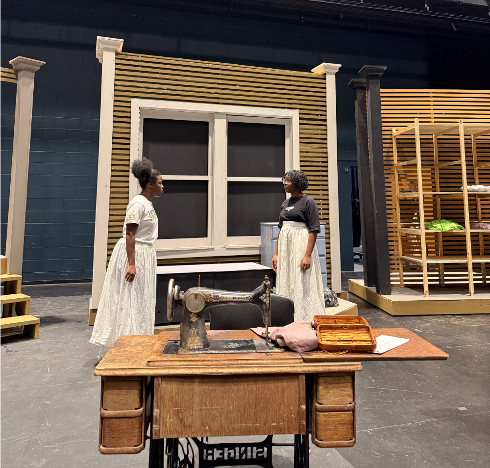 two cast members in dresses stand on stage, a sewing machine in a desk is in the foreground