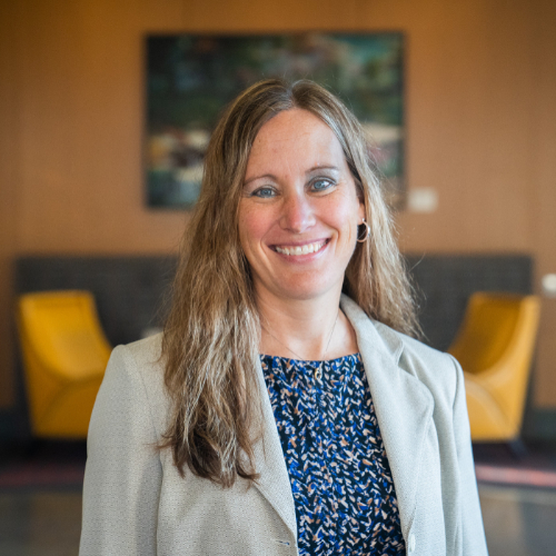 Erica Hamilton portrait, standing in a room wearing light jacket over blue print shirt