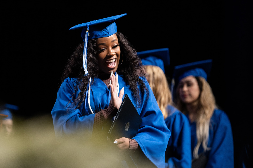 graduate in cap and gown walking across stage, waving to audience, two students in gowns behind first person