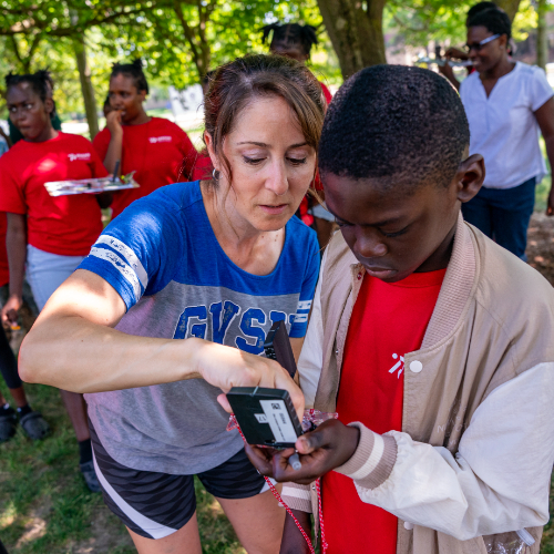 Faculty member in GVSU t-shirt shows young student in red shirt and light jacket how to use a compass outside