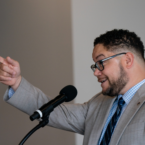 Danny Velez points to the audience while standing at a podium with a microphone