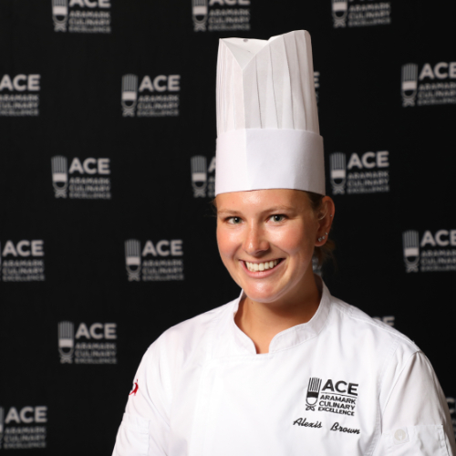 Chef Alexis Brown in chef hat and white coach standing in front of ACE backdrop