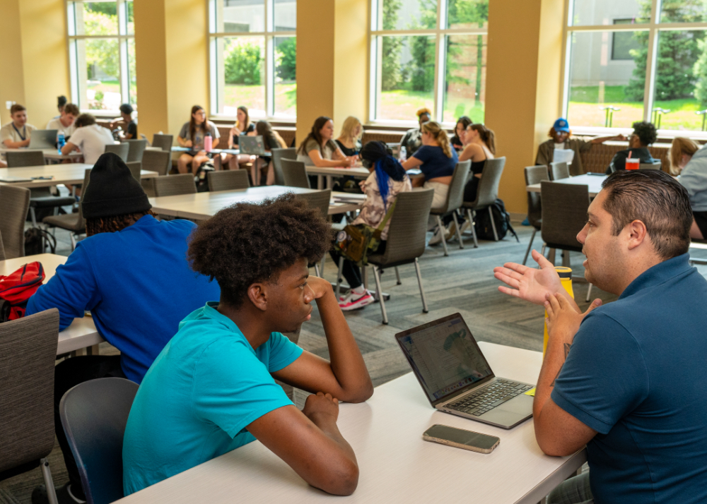 two people sit across a table with a laptop in front of the man on right; other students seated at long tables in the room