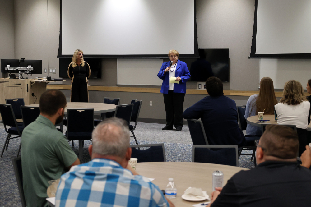 two women standing at front of room, women on right in blue blazer with microphone