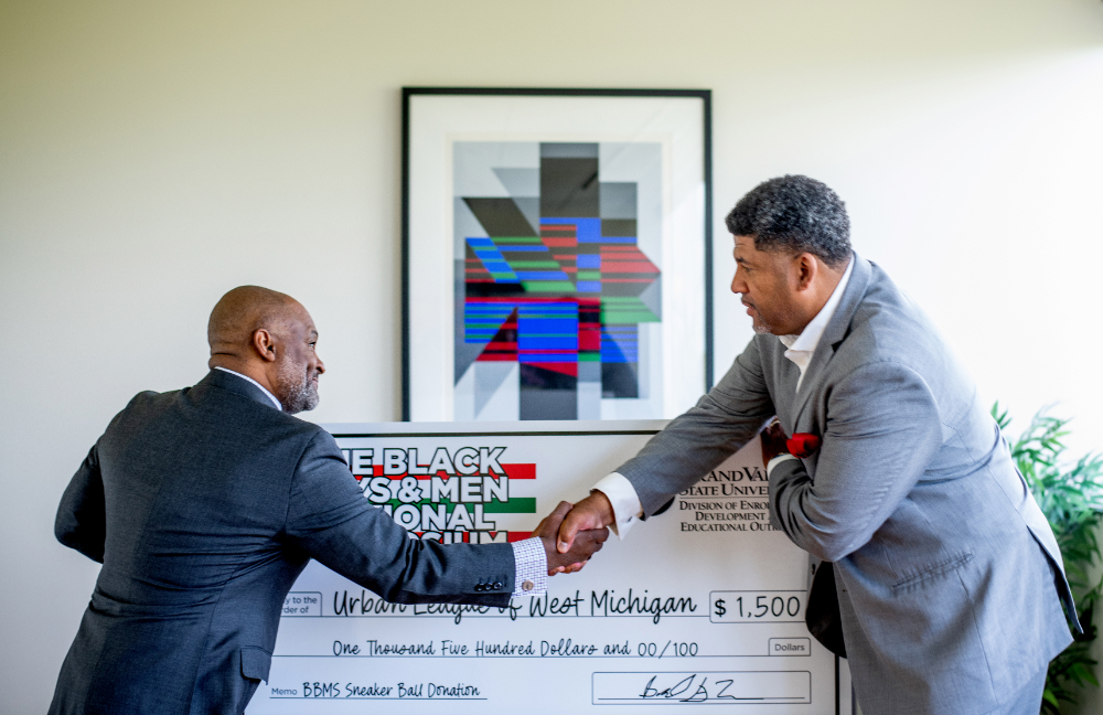 two men shake hands in front of oversized check from the Black Boys and Men Symposium