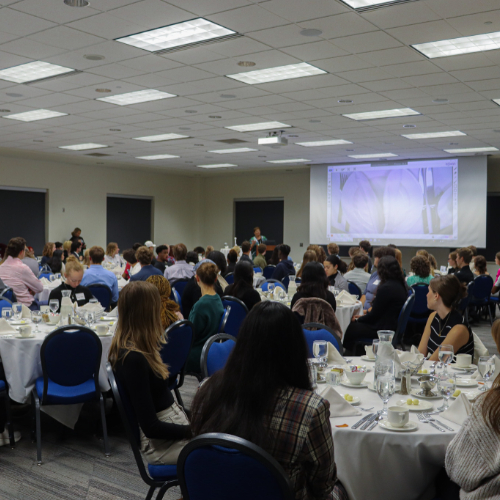 Cindy Brown up front at podium, projection screen shows place setting, students seated at round tables