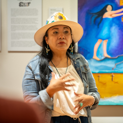 Reyna Garcia in hat with sunflower stands in front of colorful painting