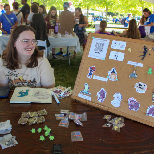 student sits in front of table with stickers she made, a cork board displays more