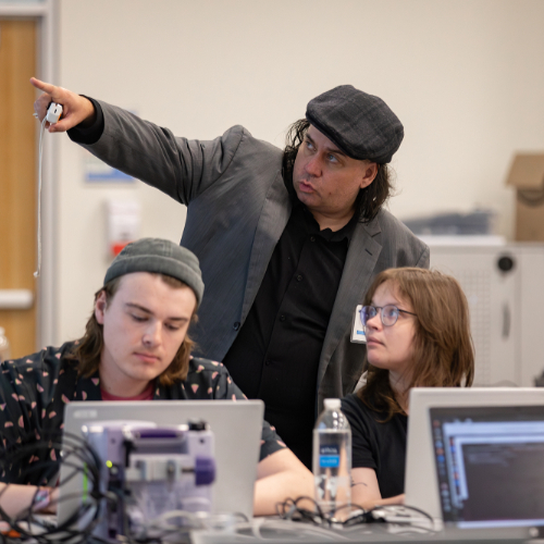 instructor standing and pointing, three students on laptops looking where instructor, in hat and suit jacket, is pointing