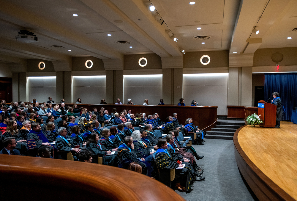 faculty member behind podium on stage, audience of faculty in academic regalia seated in rows