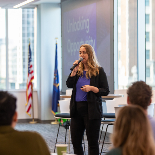 Rachel Salinas with microphone standing to speak to audience at tables. Projector screen reads Unlocking Potential and there are US and Michigan flags standing at left