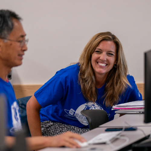 two people in blue t-shirts in computer lab, woman at right is smiling facing the camera