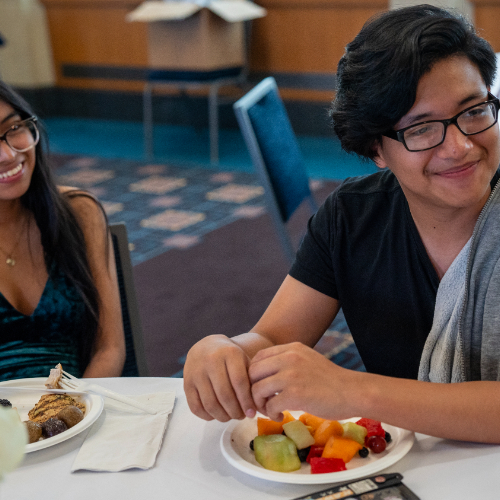 two students seated at table smiling, plates of food in front of them