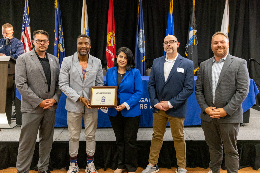 five people standing in front of row of military flags, two people sharing the hold of a framed certificate