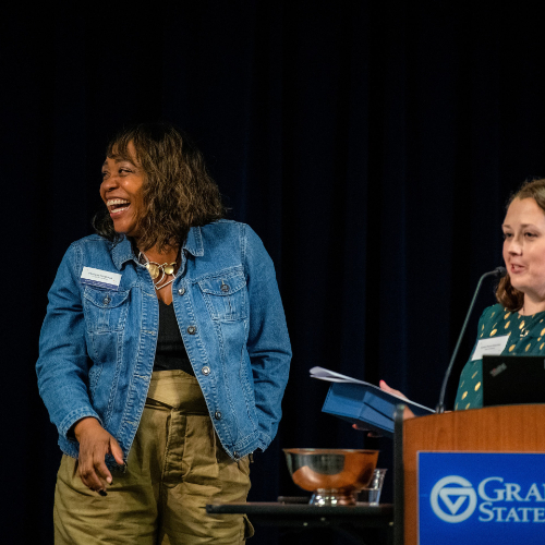 two women on stage, woman on left laughing while woman behind podium presents her with an award