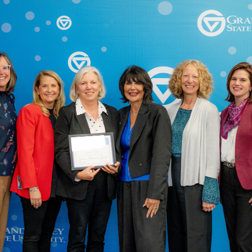 six women standing against a GVSU backdrop; woman third from left holds a certificate