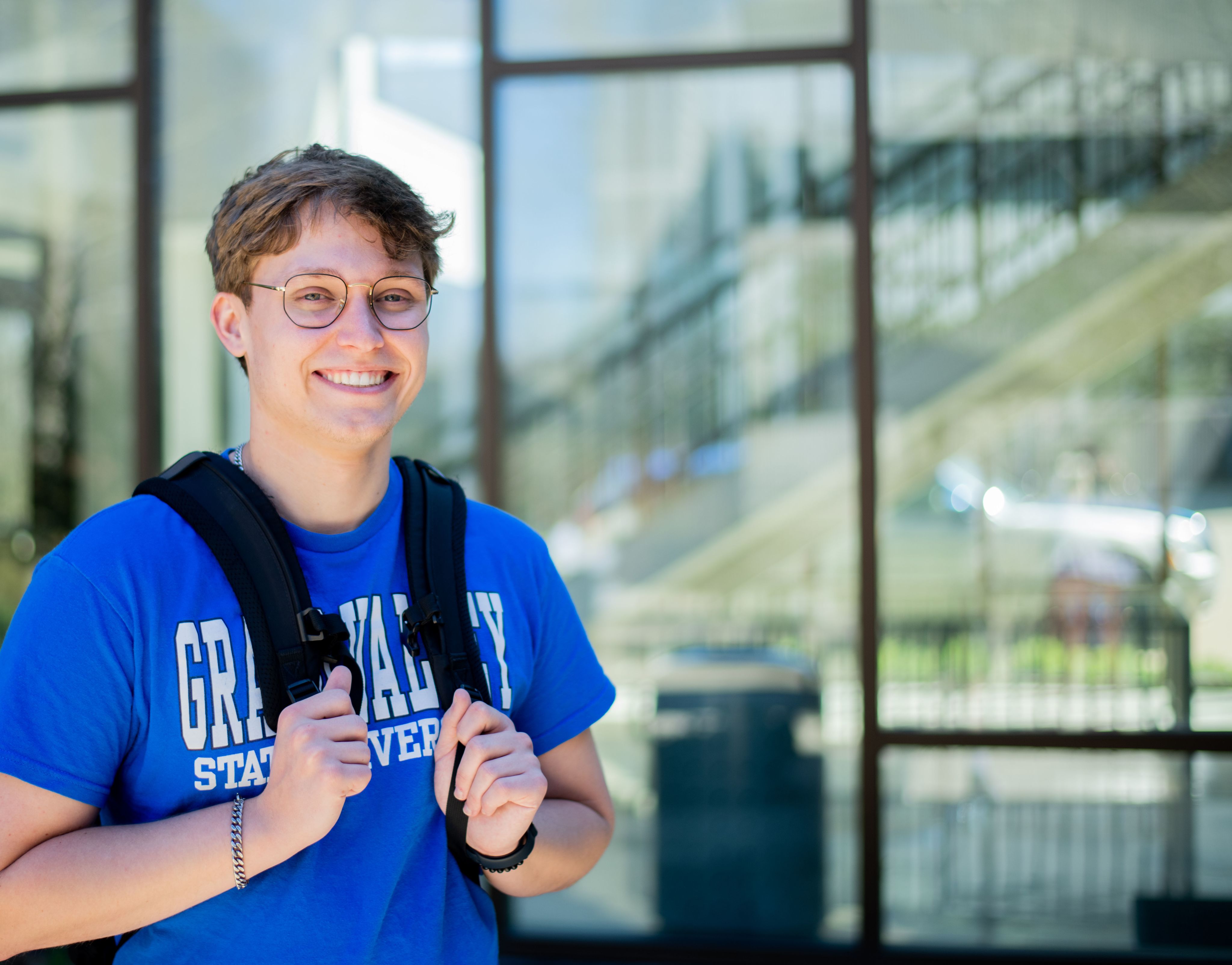 student with hands on backpack straps next to building with full windows, can see staircase inside
