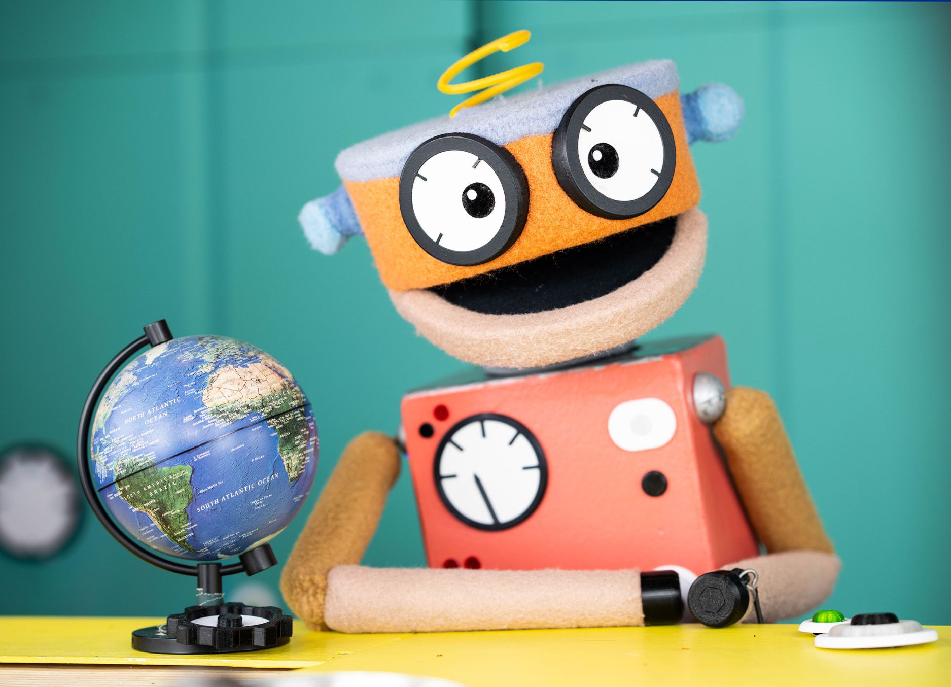 Wimee, an orange robot looking puppet sits at a desk with a small globe