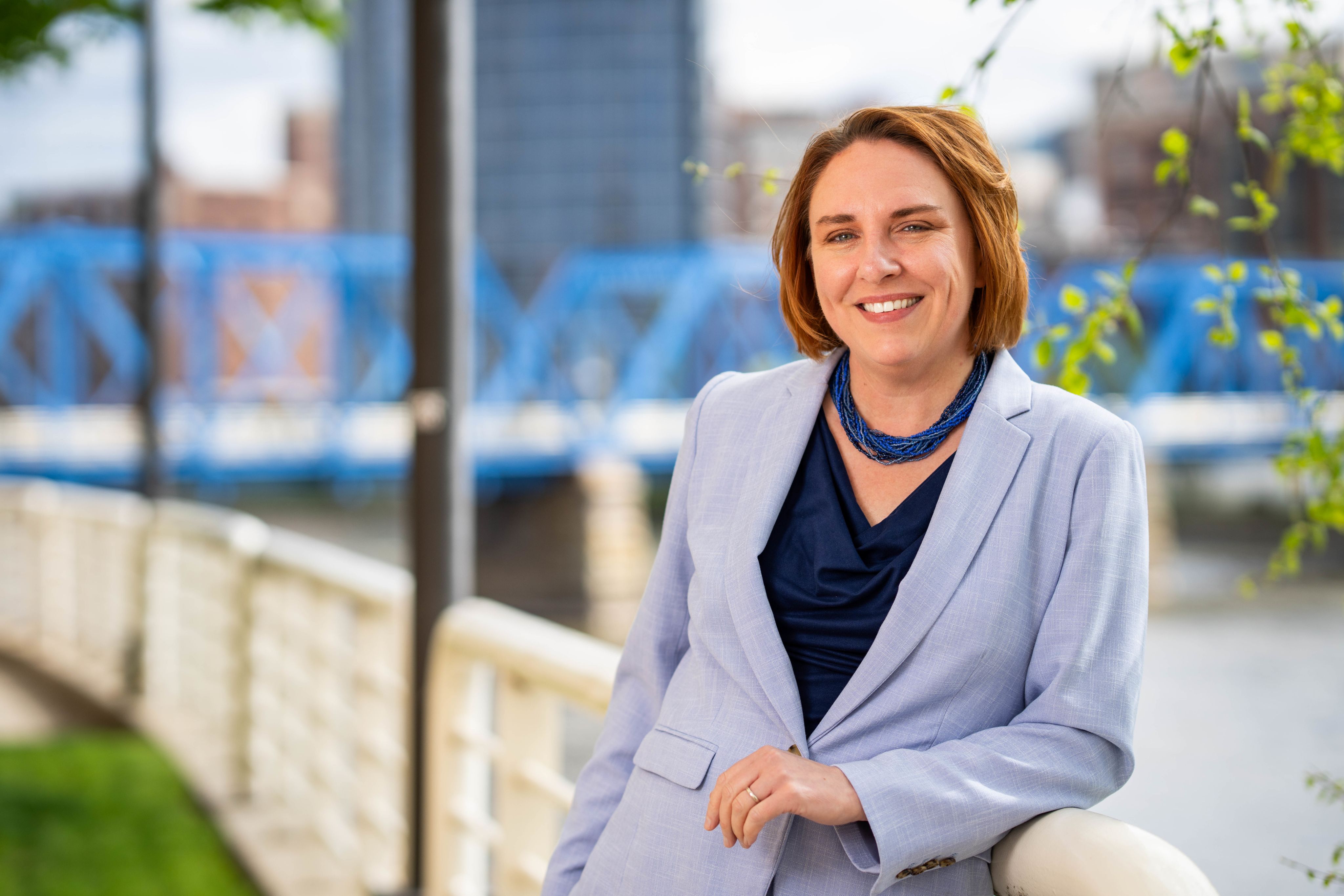 Kara Van Dam in light blue jacket and blue shirt leaning against bridge over Grand River