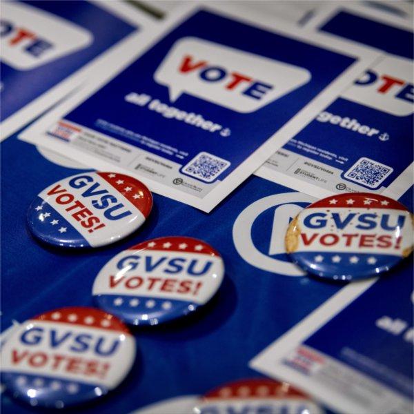 A collection of buttons that read �GVSU Votes!� in blue and red text sit on a blue Grand Valley tablecloth. Behind them are flyers with a QR code that say �Vote! All together�.