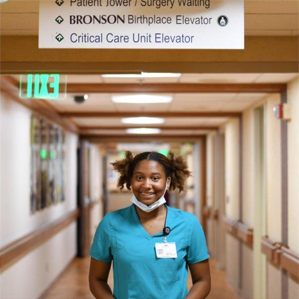 Mariah Rudolph stands in hospital hallway in blue scrubs and face mask pulled down to chin