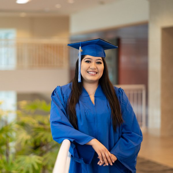A student in a cap and gown smiles while leaning against a railing.