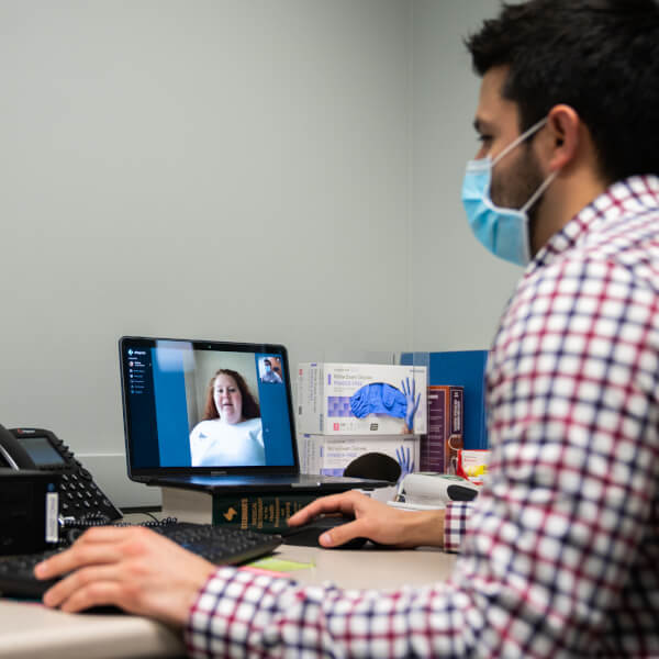 man wearing medical mask sitting at computer conducting a zoom-like meeting with a womam