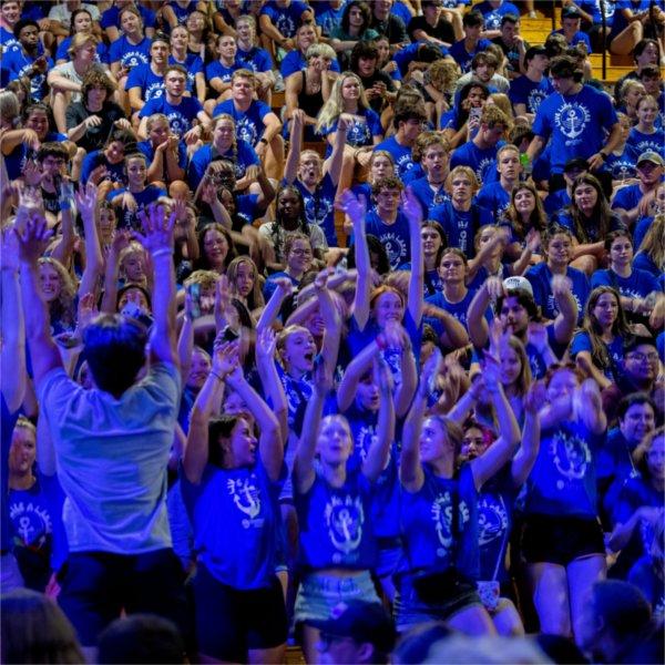 A group of students in blue Laker for a Life shirts sit in the bleachers and hold their arms up to do the wave.