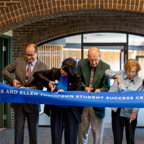 people stand behind a blue ribbon, Bob and Ellen Thompson Student Success Center, with scissors in hand to cut the ribbon