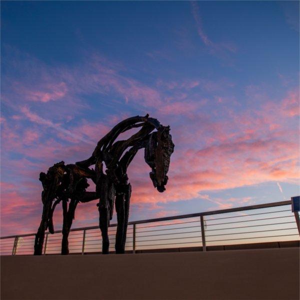 A horse sculpture is seen along a railing against a pink sky.