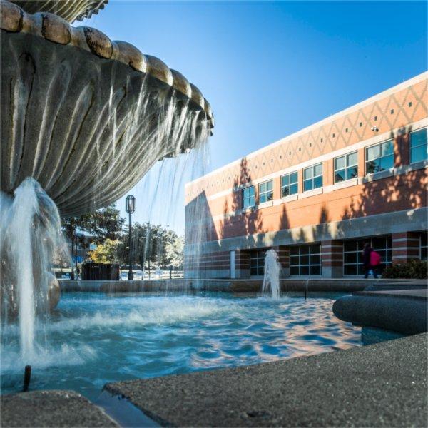 A water fountain sprays water on a sunny day. The Student Services building can be seen behind it.