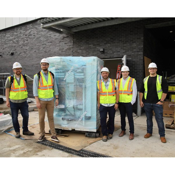group of five people standing outside in construction yellow vests, with a large, wrapped cyclotron in center