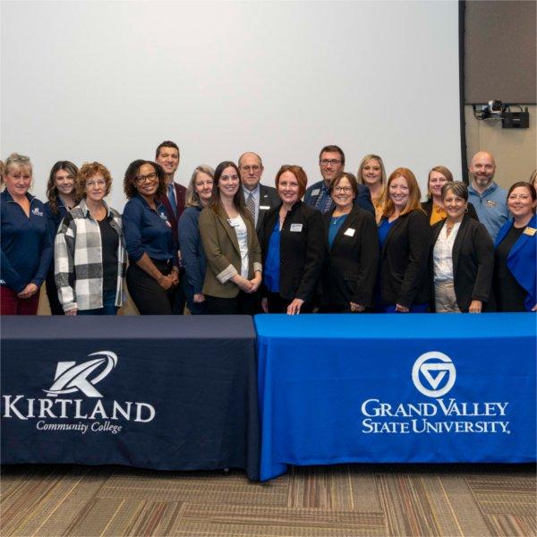 large group of people standing in two rows behind 2 tables draped with banners for GVSU and Kirtland Community College