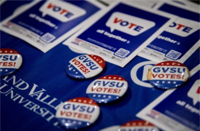 A collection of buttons that read &ldquo;GVSU Votes!&rdquo; in blue and red text sit on a blue Grand Valley tablecloth. Behind them are flyers with a QR code that say &ldquo;Vote! All together&rdquo;.