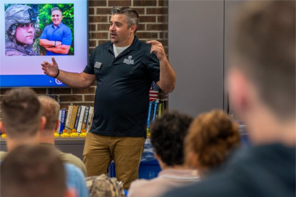 A person gestures while speaking before a group. There are pictues of the person on a screen in the background.