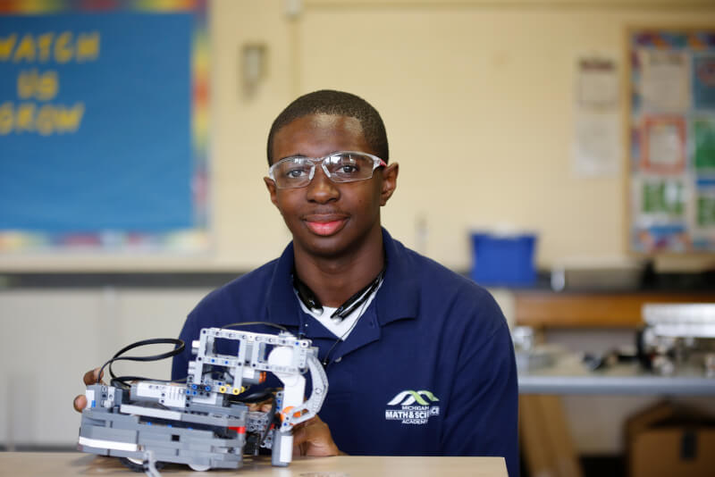 A student at Michigan Mathematics and Science Academy poses for a photograph with a robotic device while seated at a desk in a classroom.