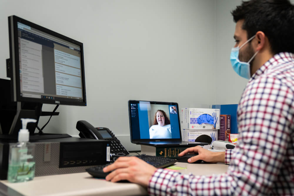 man wearing medical mask sitting at computer conducting a zoom-like meeting with a womam
